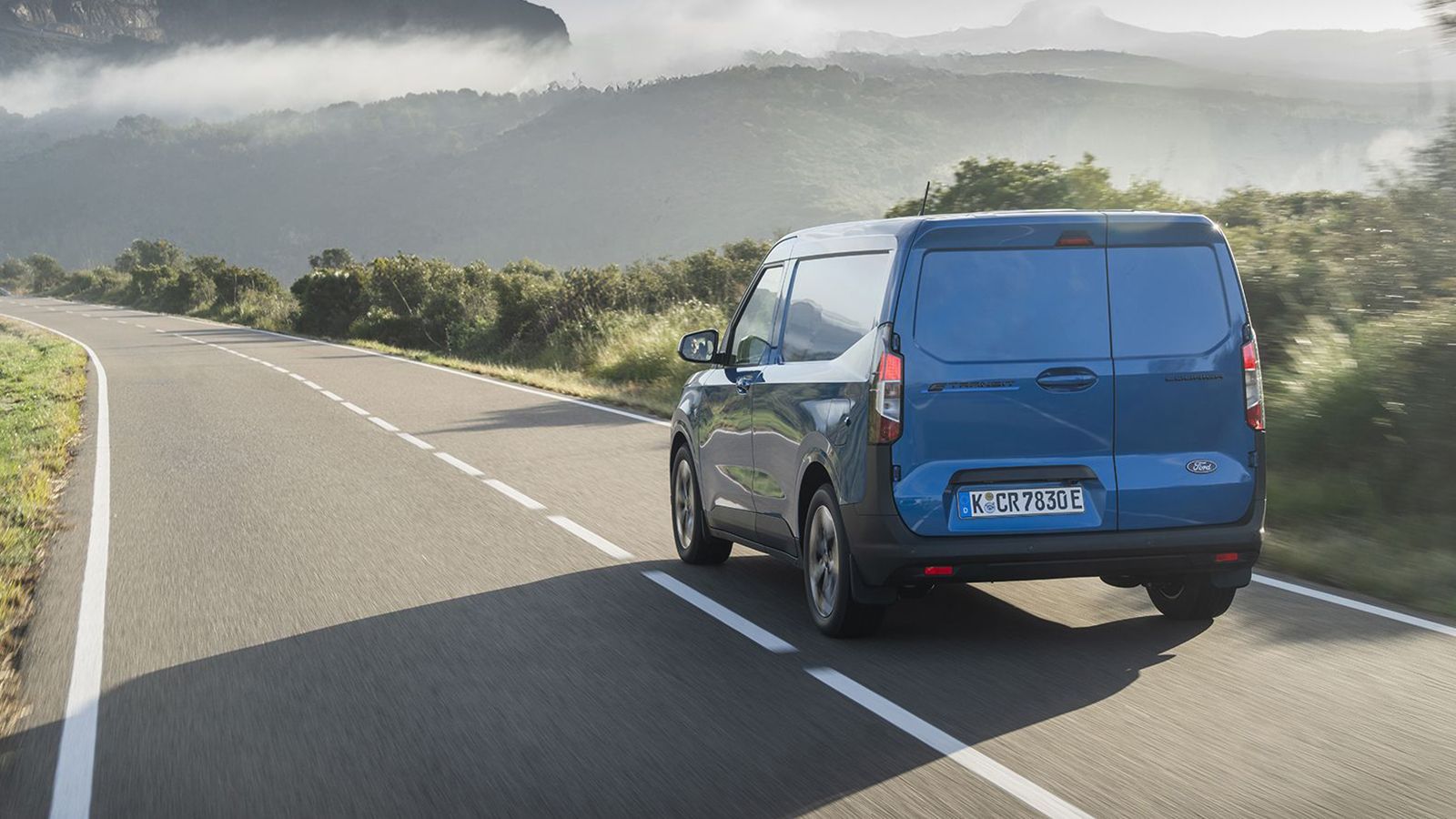 Μπλε Ford E-Transit Courier σε πίσω πλάγια λήψη σε επαρχιακό δρόμο.