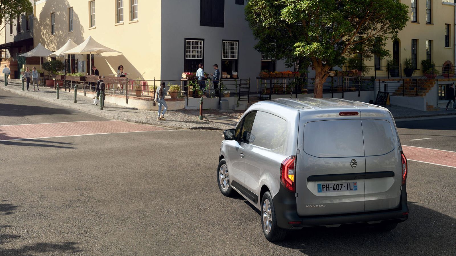 Ασημί Renault Kangoo Van από πίσω και πλάγια σε αστικό δρόμο, μπροστά από καταστήματα.