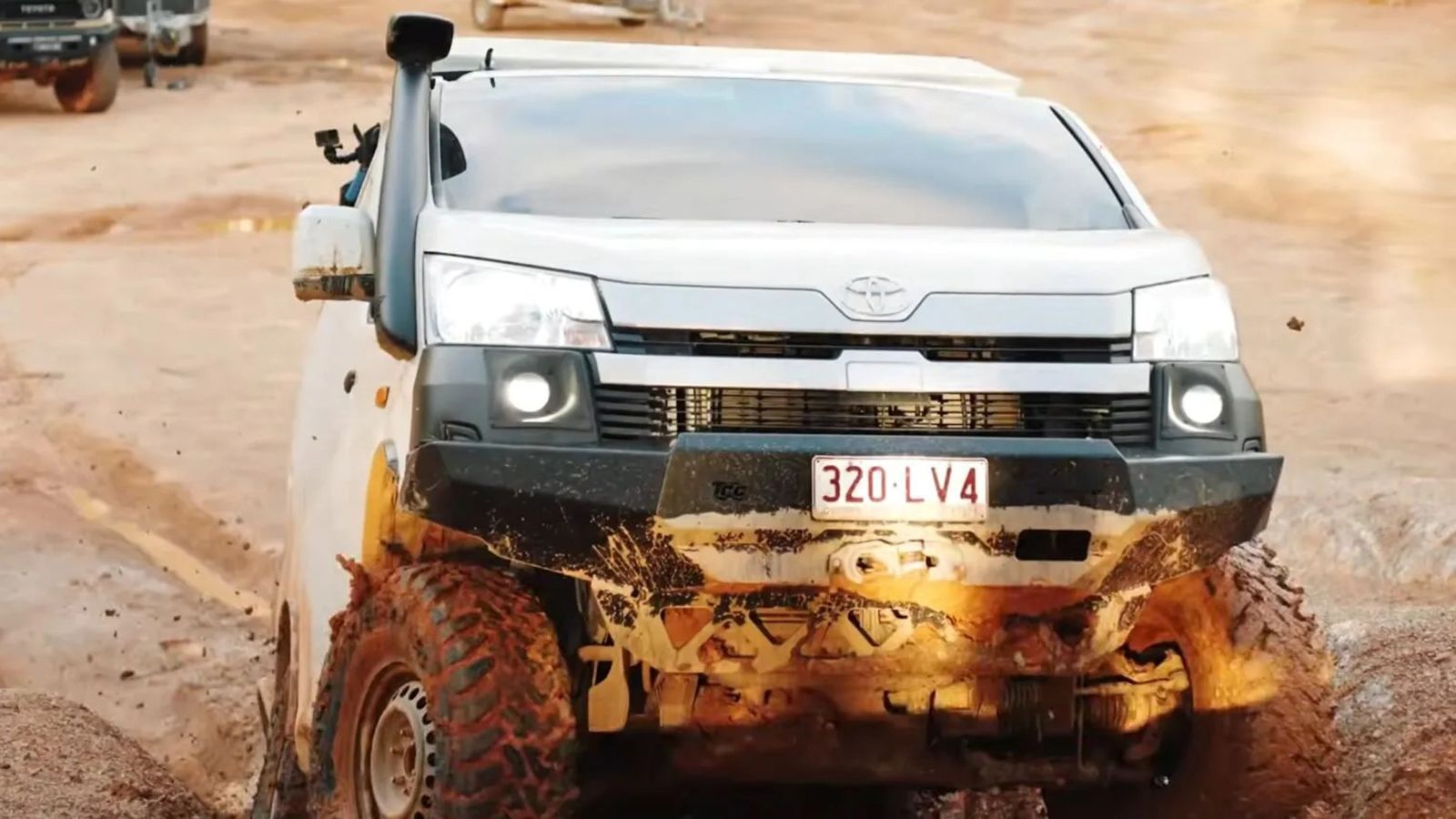 Λευκό Toyota HiAce 4x4 κινείται σε βαθιά αυλάκια άμμου ρυμουλκώντας σκάφος.