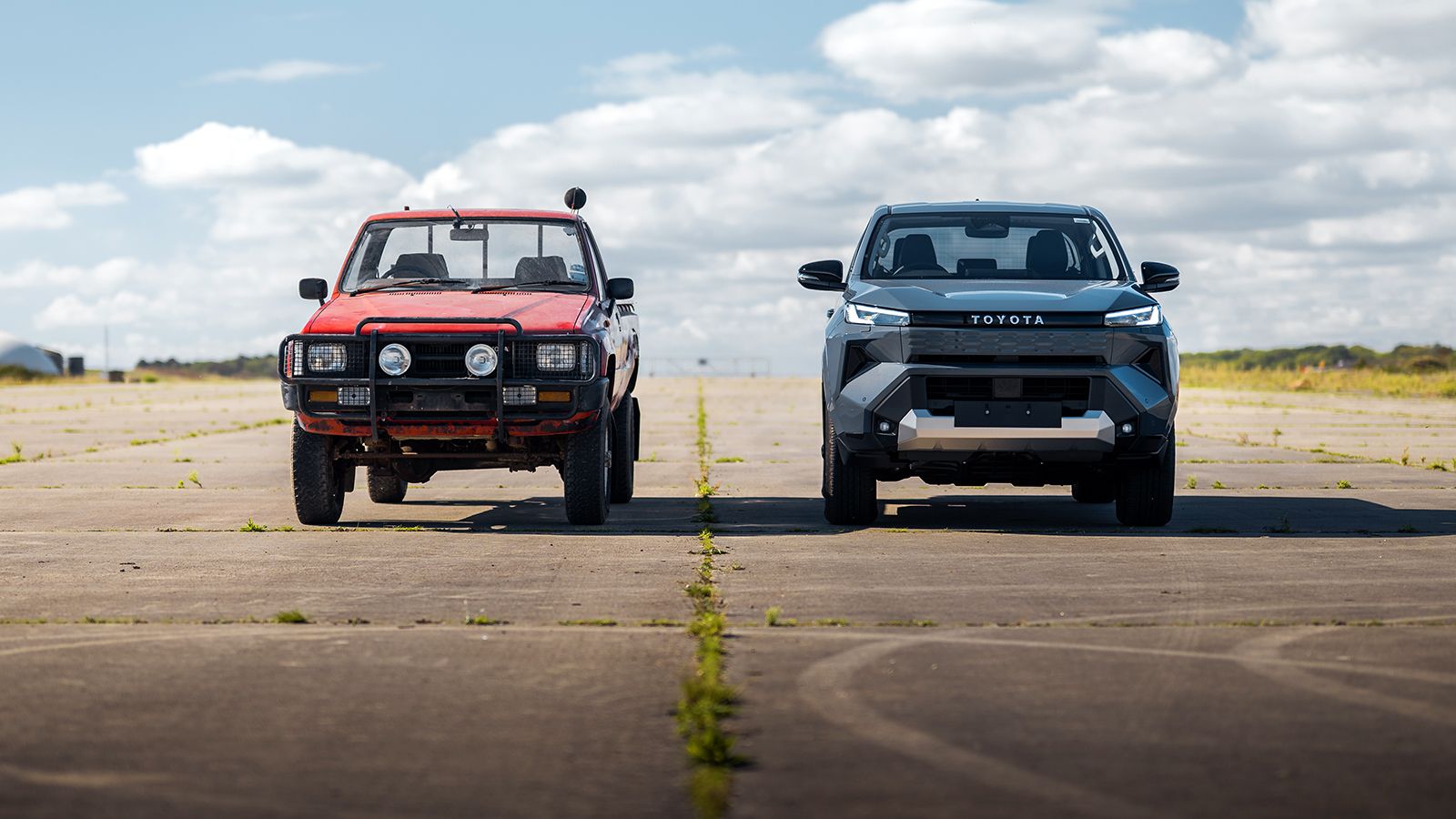 Παλιό κόκκινο Toyota Hilux δίπλα στο νέο ηλεκτρικό Toyota Hilux σε πίστα αεροδρομίου.